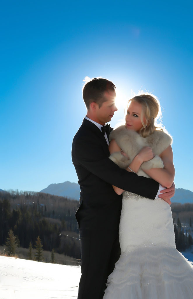 couple poses with the sun behind her, bride clutches her fur in the snow.